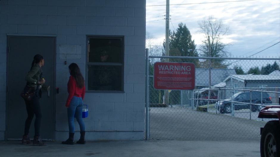 Maddie opens the door leading into the lab for Ryn while in the distance Ian Sutton watches from his car.