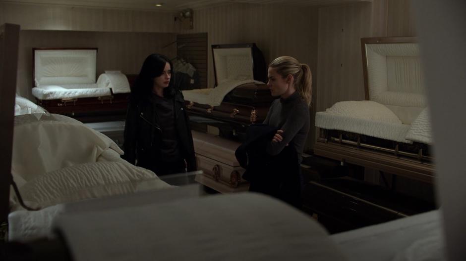 Jessica approaches Trish who is standing in the coffin showroom.