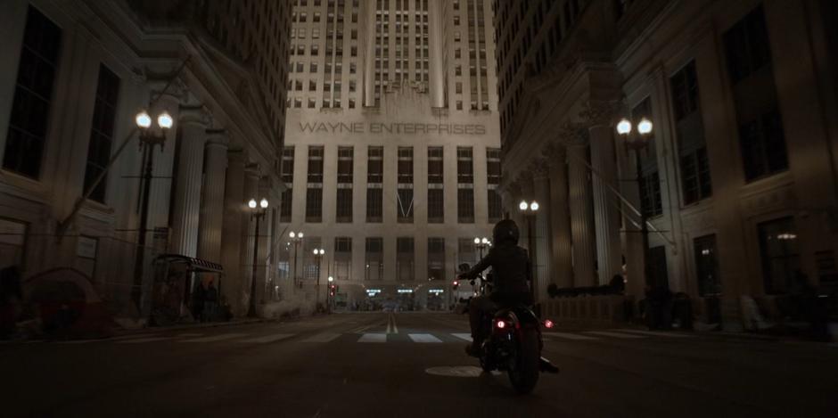 Kate rides her motorcycle down the street towards the Wayne Enterprises building.