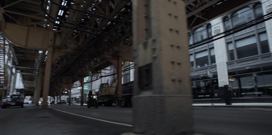 Kate drives her motorcycle down the street under the elevated rail.