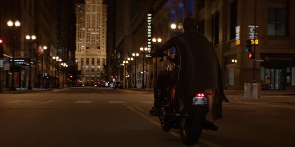 Kate rides her motorcycle down the street leading to the Wayne Enterprises building while wearing her Bat costume.