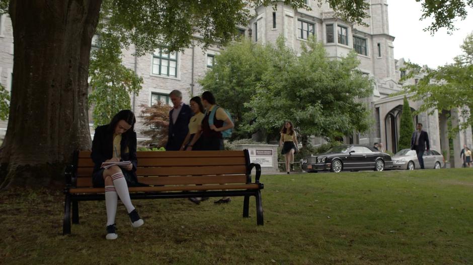 Young Lena sits alone doing homework on a bench in front of the school as Andrea approaches from the distance.