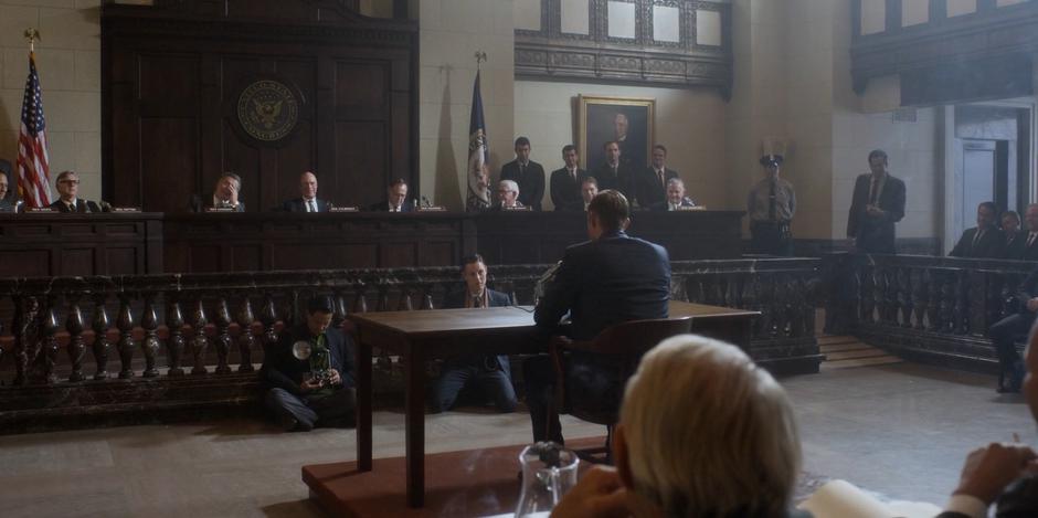 Ed sits at the table in the middle of the room while Representative Sandman questions him.