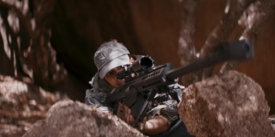 A sniper lies on some rocks looking through her scope.