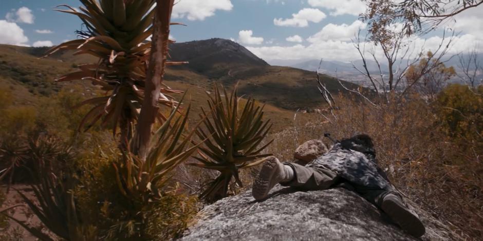 The sniper lays on a rock preparing to fire at the truck in the distance.