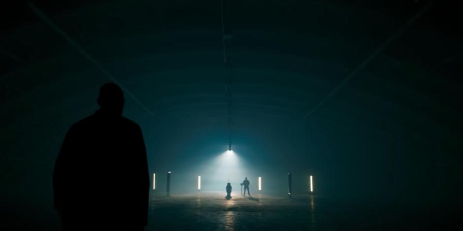 Daniel Barton walks through the dark hanger to where a guard is watching over his mother in a pool of light.