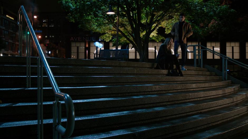 Barry walks over to where Sara is sitting on the steps in the plaza at night.