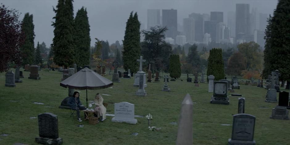 Alice pours tea for Mouse and herself as they sit under an umbrella next to Catherine's grave in the rain.