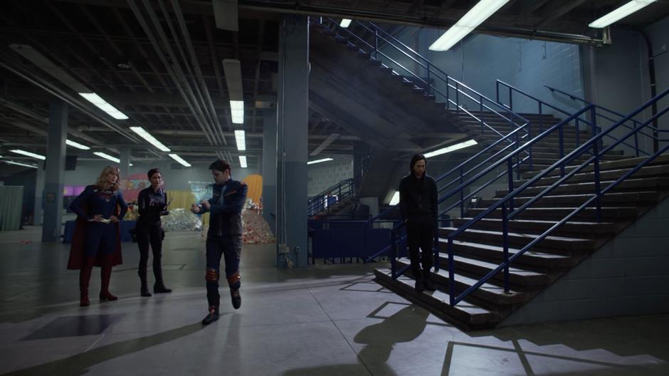 Kara and Alex talk with Winn who is scanning the building while Brainy leans against the railing on the stairs.