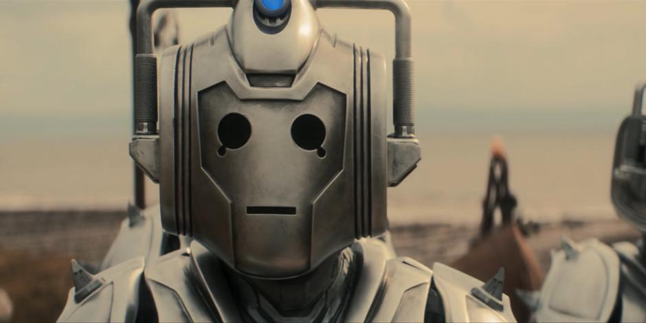 A Cyberman walks down the path towards the camp.