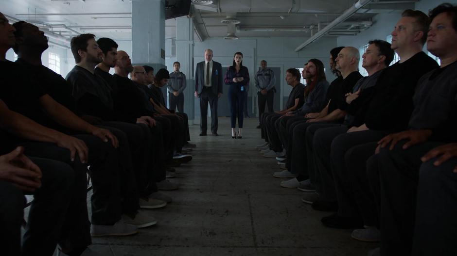 The warden and several guards watch as Lena activates the program on a group of prisoners seated in two lines.
