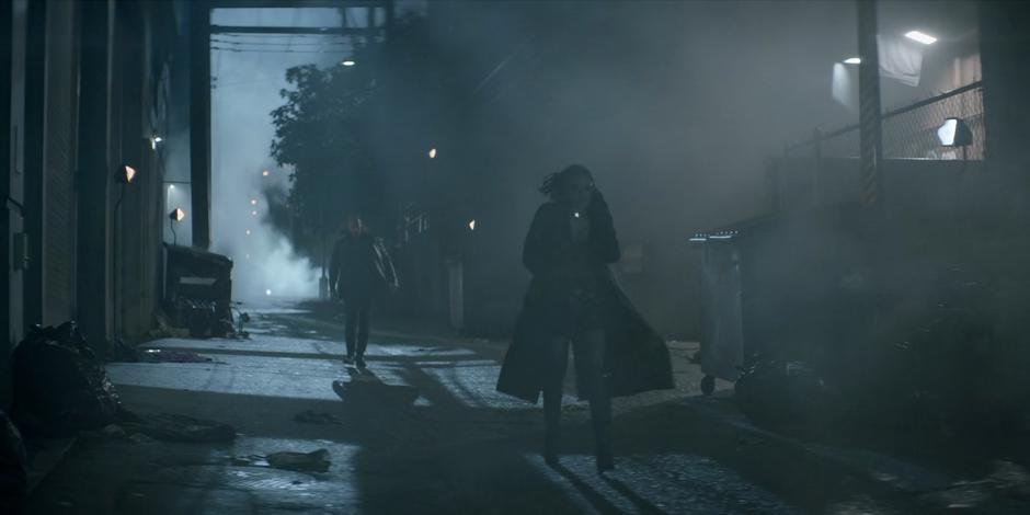 A woman walks down a dark and foggy alley talking to her friend on the phone while a man follows her.