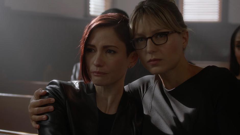 Kara pulls Alex against her shoulder as they listen to the eulogy.