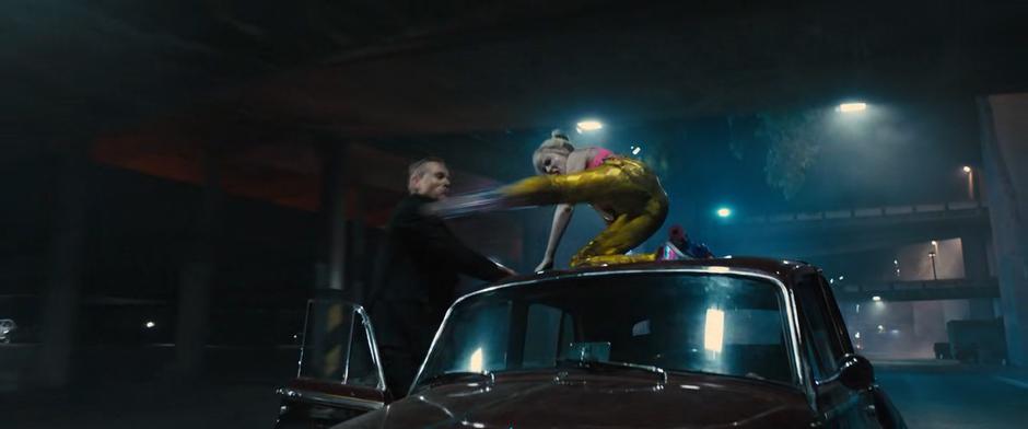 Harley kicks one of the goons while kneeling on the roof of Roman Sionis's car.