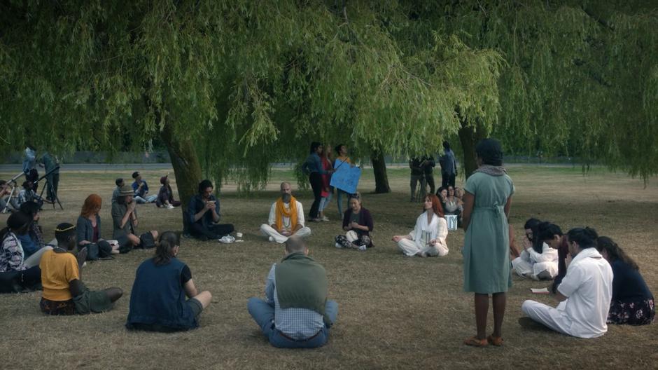 Eliot sits against a tree eating his sandwich while people pray in the circle around him.