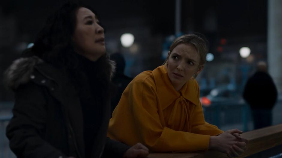 Villanelle looks at Eve while leaning up against the railing next to her.