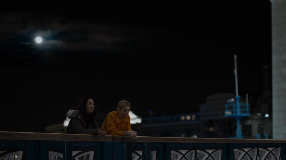 Eve and Villanelle lean on the edge of the bridge looking out over the river.