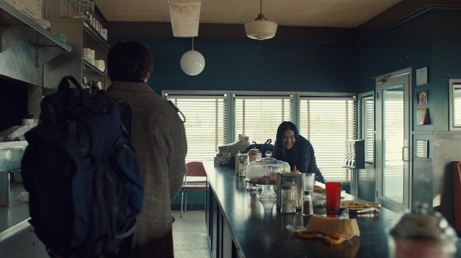 Rachel sits at the diner while Billy walks towards her from behind the counter.