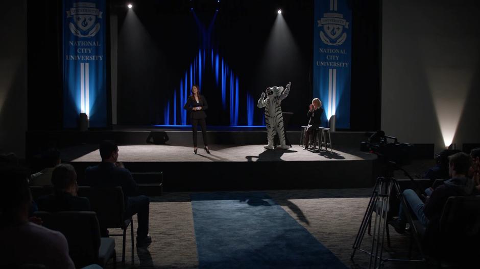 Andrea announces her new VR project while the school's mascot cheers her on from next to her on stage.