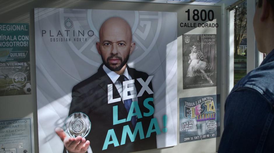 Two people stand in front of a Lex Luthor poster in spanish.