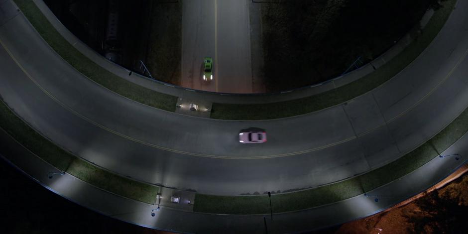 The pink car drives over the overpass while the green one drives beneath.