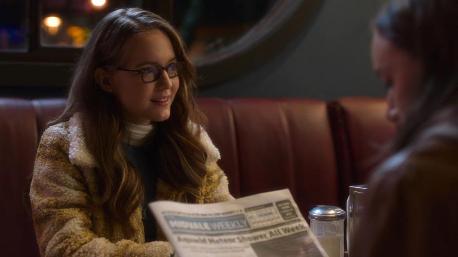 Young Kara stares after Kenny when he leaves the table as Alex looks back at the newspaper.