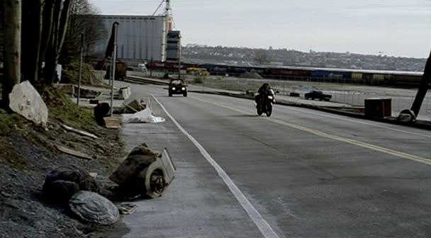 Max rides her motorcycle down a road by the port.