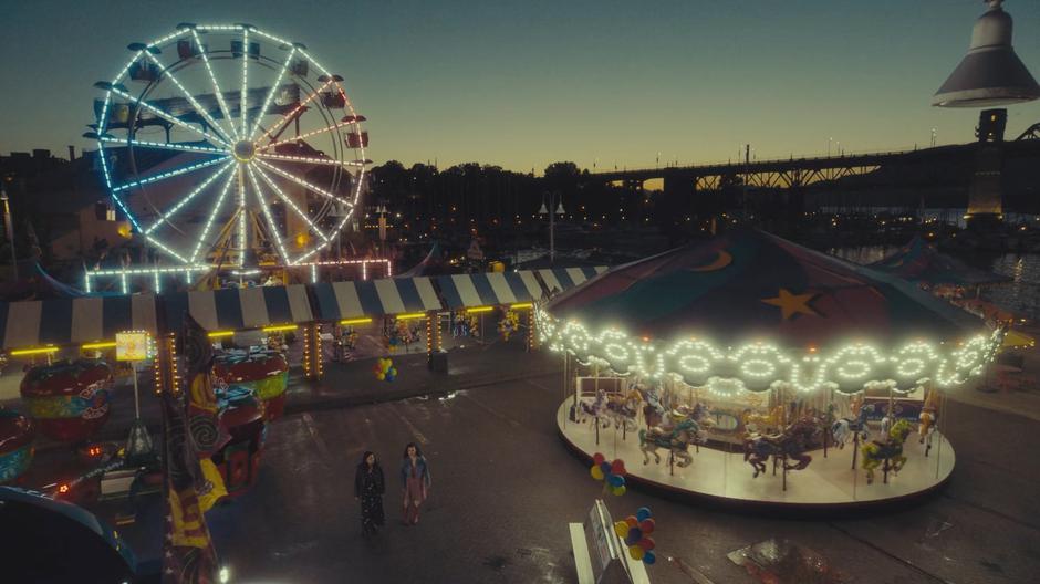 Mel and Magie stand in the middle of the empty carnival next to the carousel.