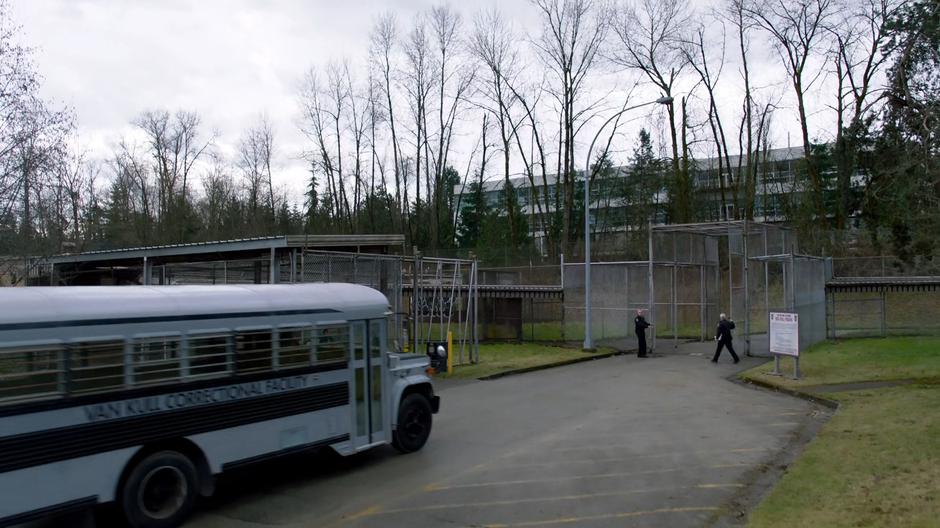 Two guards open the gate as a blue prison transport bus approaches.