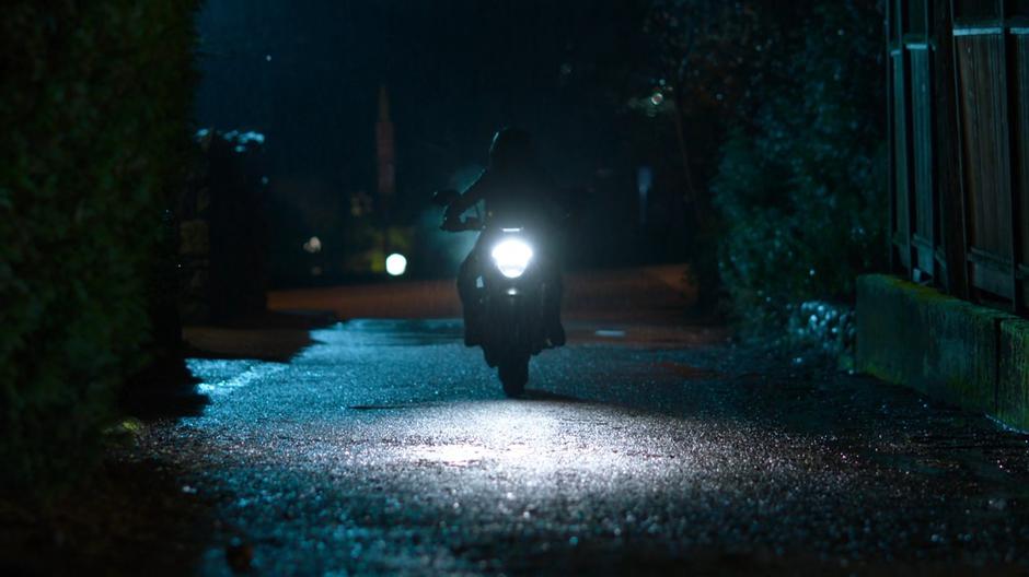 Kelly drives into the alley behind the house on her motorcycle at night.