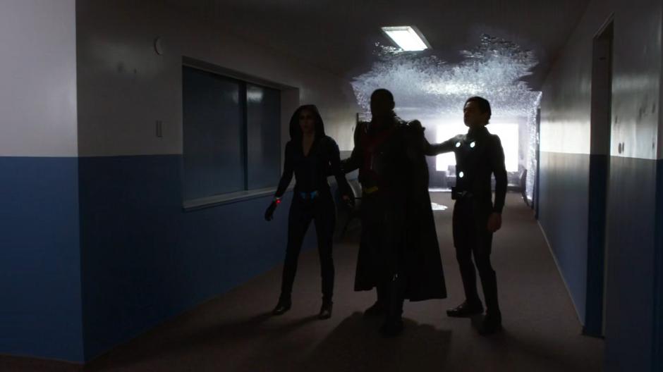 Alex, J'onn, and Brainy phase through the floor into the hallway as the ice approaches from behind them.