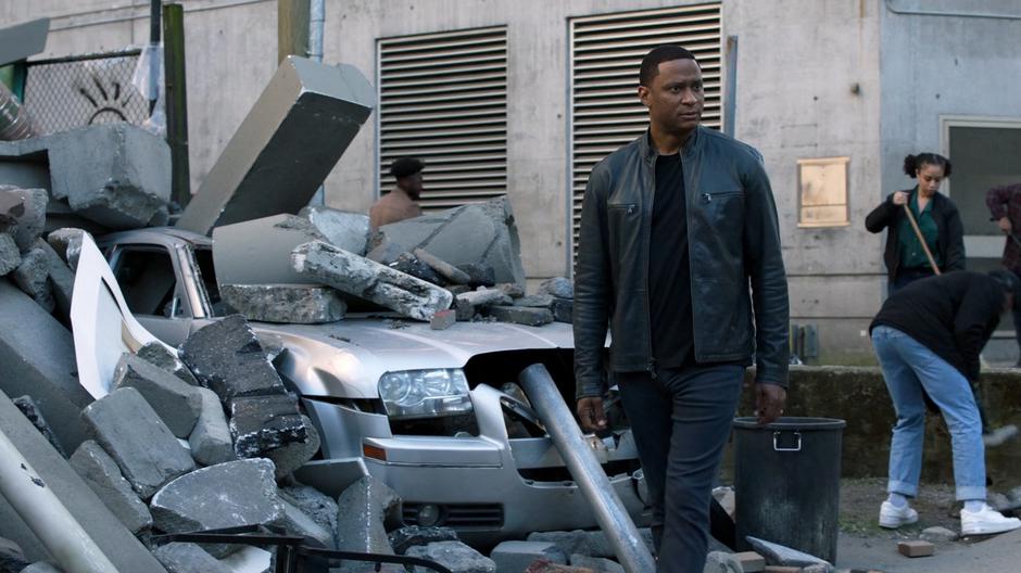 Diggle looks around the ruins of the building while people clean up in the background.