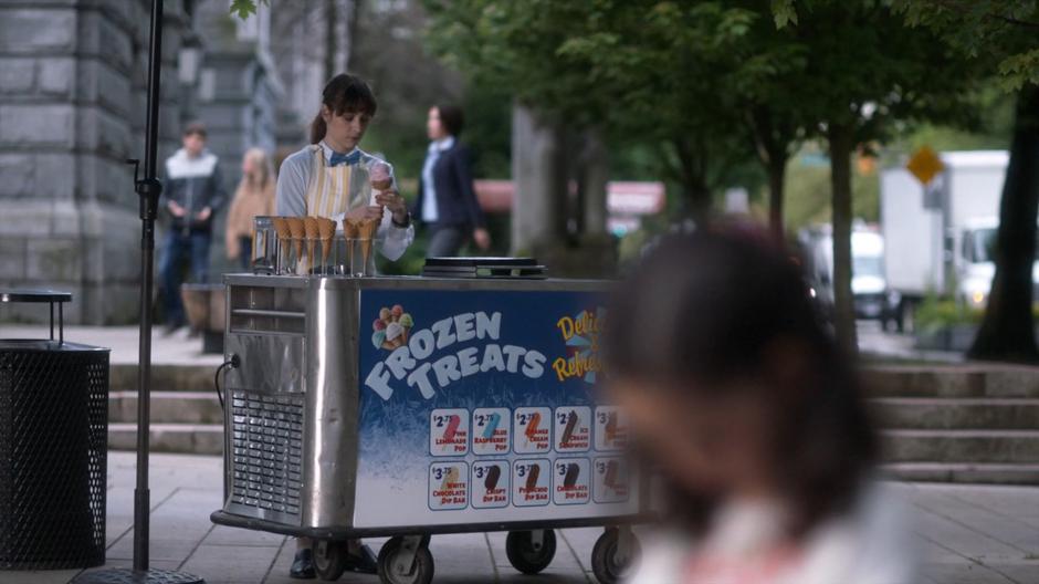 A woman running an ice cream cart prepares another cone as a young girl in the foreground mourns her dropped cone.