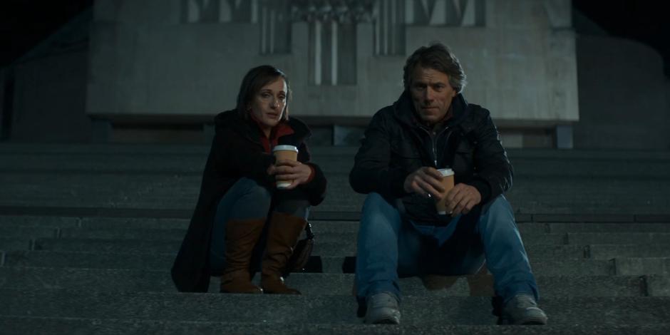 Diane and Dan sit on the steps of the cathedral at night holding their same coffee cups.