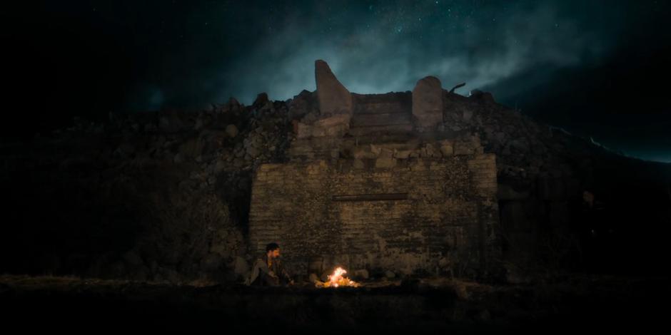 Namaca sits by his fire in the empty ruins at night.