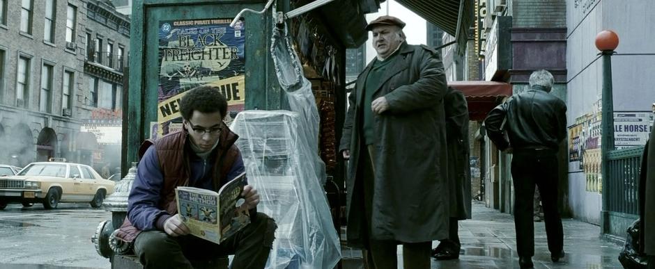 A kid reads his comic book by the news stand.