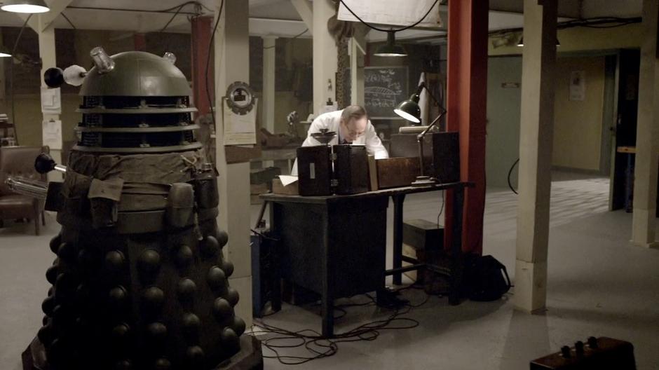 Bracewell goes over some notes while a Dalek drives past.