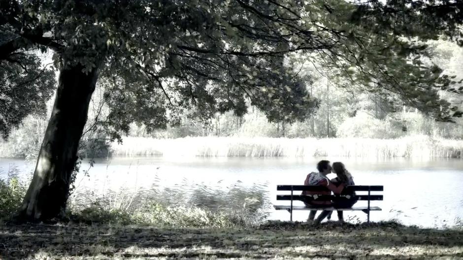 Amy and Rory kiss on a park bench in front of a lake.