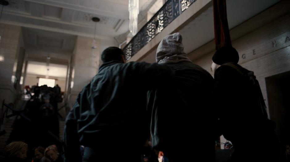 Two of Bane's thugs drag a hooded Bruce into the courthouse foyer.