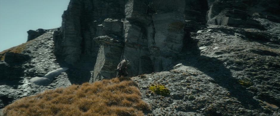 Gandalf climbs a hillside towards a steep rock face.