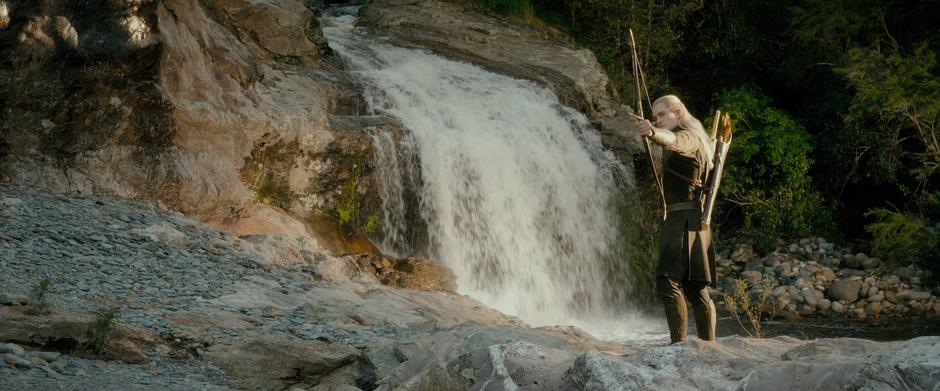 Legolas stands in front of a waterfall and points his bow at Tauriel.