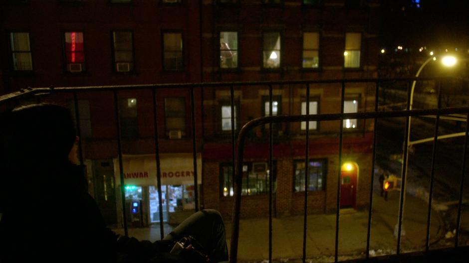 Jessica sits on the fire escape and watches the bar across the street.