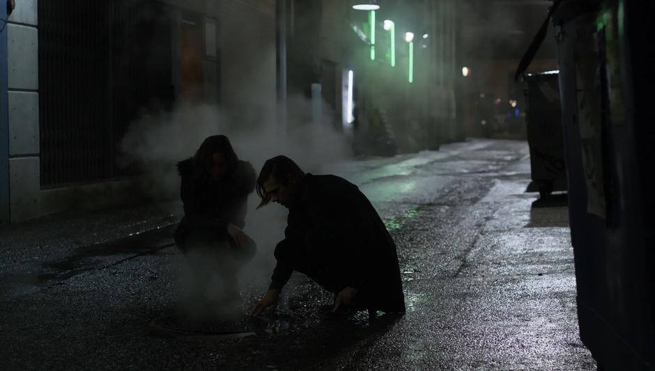 Quentin & Julia crouch down while examining the manhole cover in the middle of the alley.