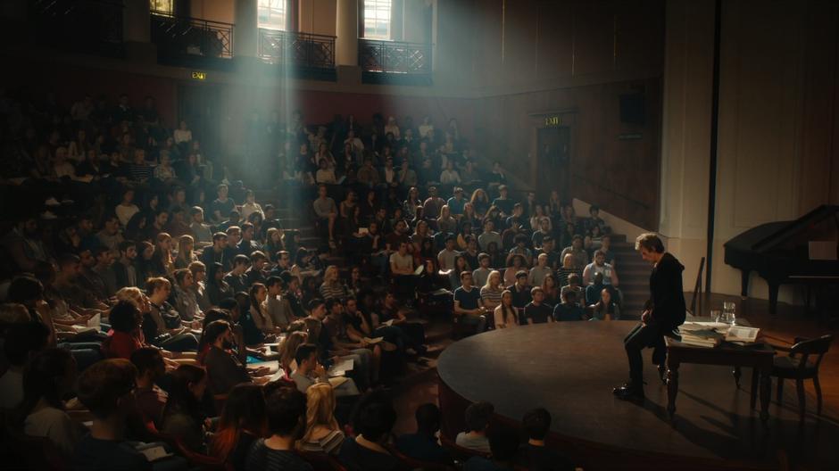 The Doctor sits on his desk in front of a hall full of students.