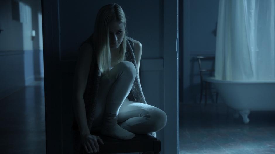 Alice sits on a chair by the door to her room after Quentin enters.