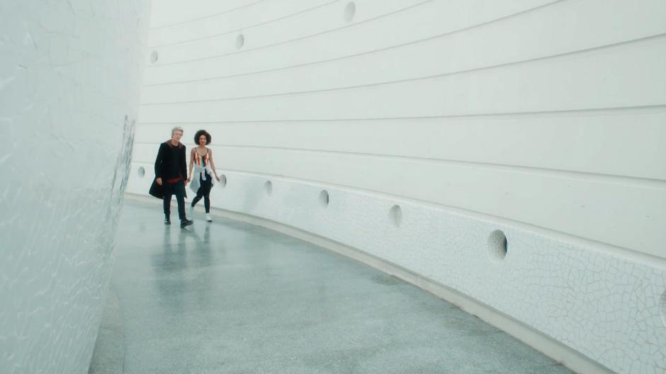 The Doctor and Bill walk around a curved corridor near the base of a large sphere.