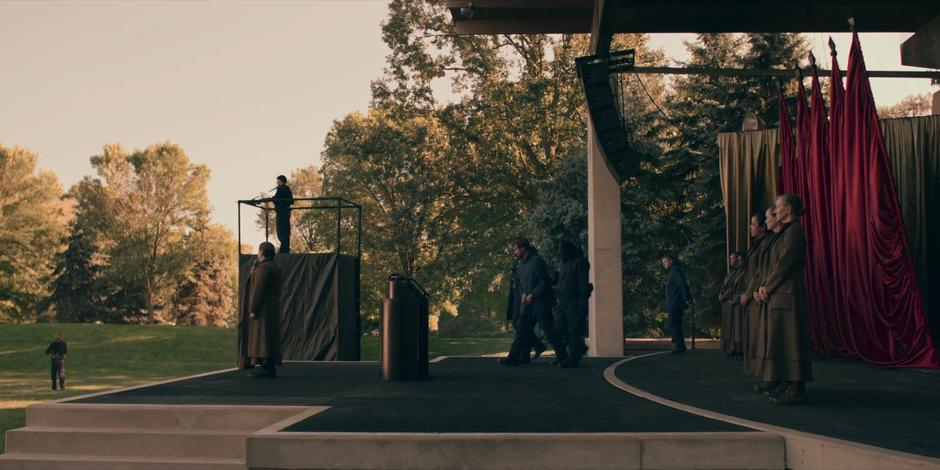 A pair of guards lead the accused onto the stage while Aunt Lydia tells the handmaids about his crime.