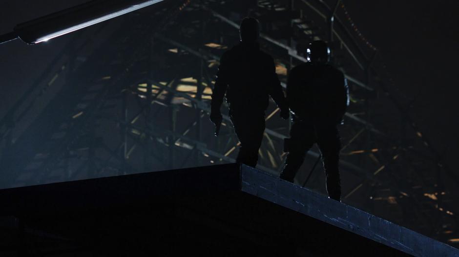 Oliver and Diggle look down on the arcade from a nearby rooftop with a roller coaster in the background.