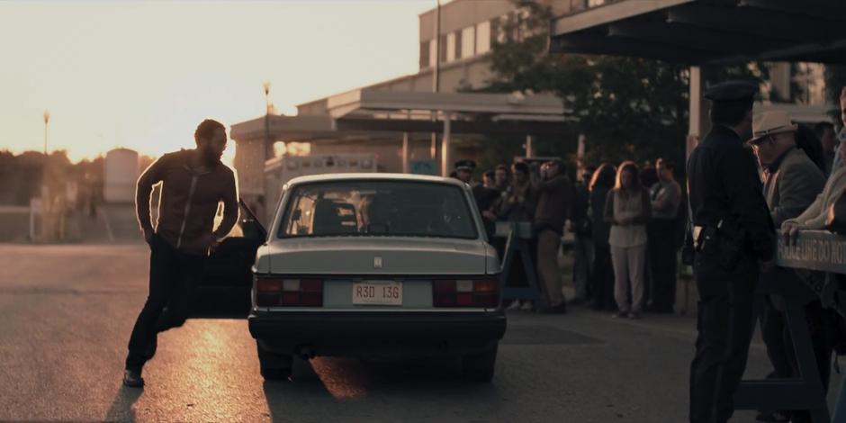 Luke runs around his car to help Offred out while the police hold back the protestors.