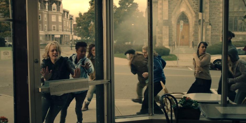 June and Moira push against the door of the cafe while protestors run past behind them.